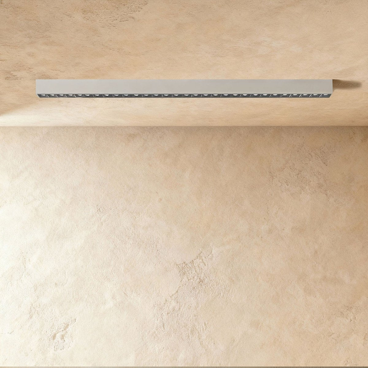 A pale silver-white linear LED bar with a row of floodlight lenses sits flush in a ceiling corner, this Clérachic fixture unlit.
