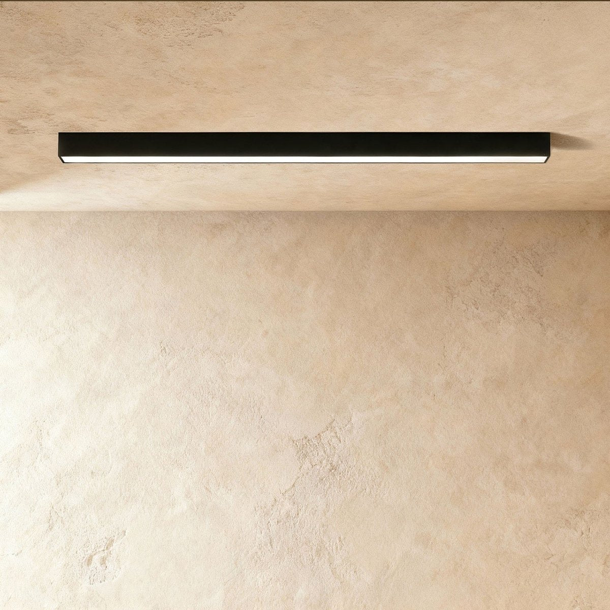 A black linear LED ceiling bar glows a soft continuous line of light in a plaster corner, this Clérachic fixture seen from below.