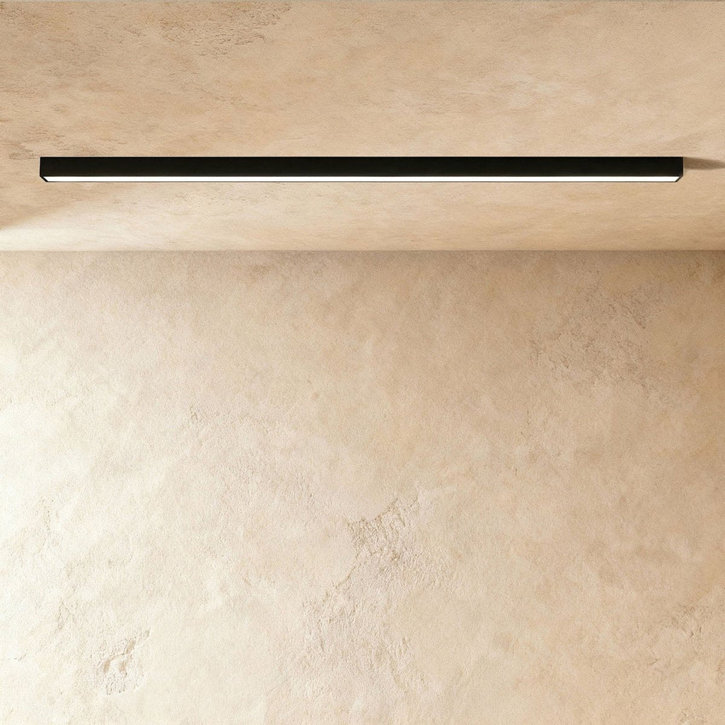 A black linear LED ceiling bar casts an even lit line across a warm plaster corner, this Clérachic fixture viewed at a slight angle.