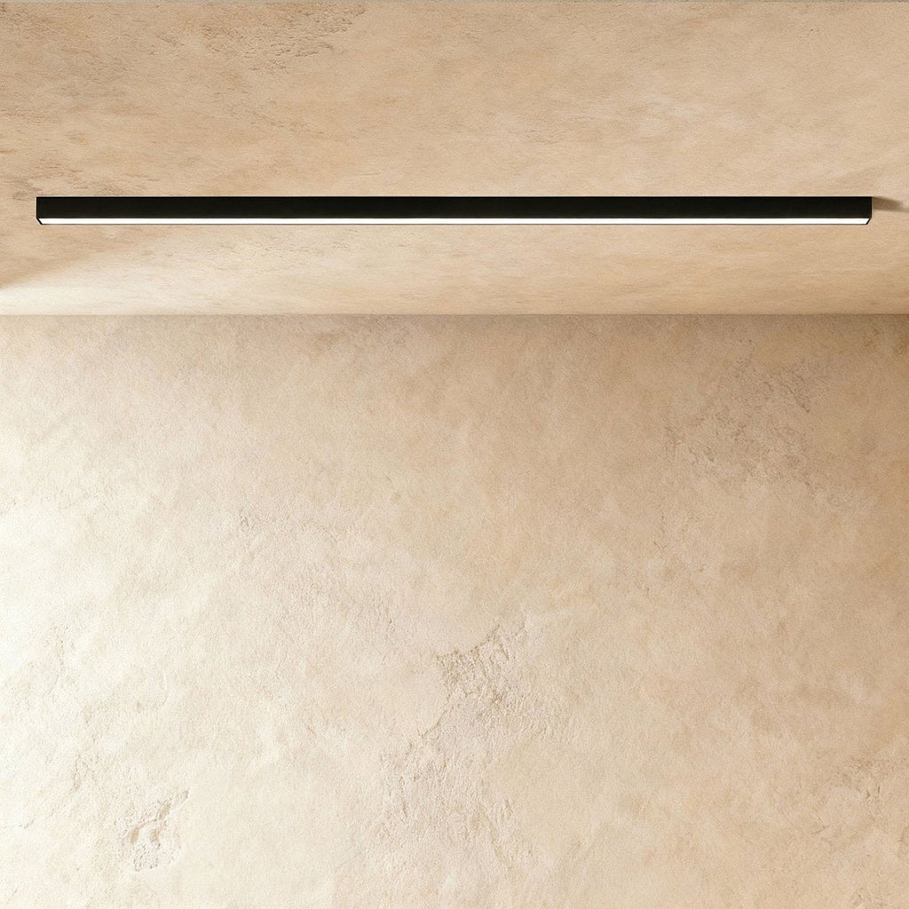 A black linear LED ceiling bar, lit edge to edge, sits flush where a plaster ceiling meets the wall, this Clérachic fixture close in view.