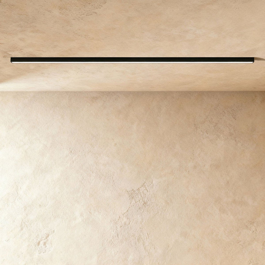 A black linear LED ceiling bar sits lit flush in a ceiling corner, this Clérachic fixture shown against a softly textured plaster wall.
