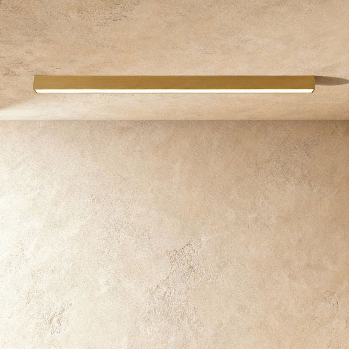 A brushed gold linear LED ceiling bar glows a warm continuous line in a plaster corner, this Clérachic fixture seen from below.