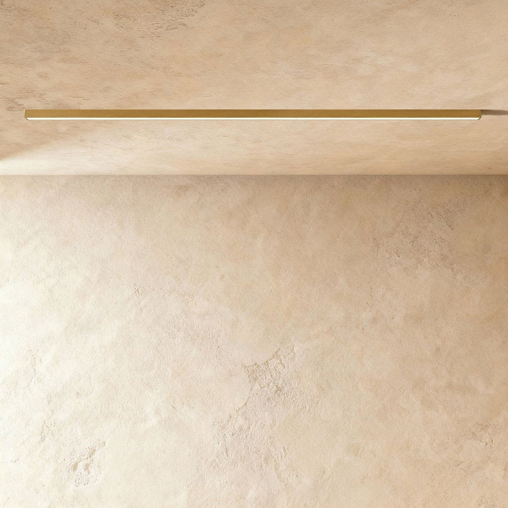 A brushed gold linear LED ceiling bar throws a warm lit line onto a plaster corner, this Clérachic fixture framed from further back.
