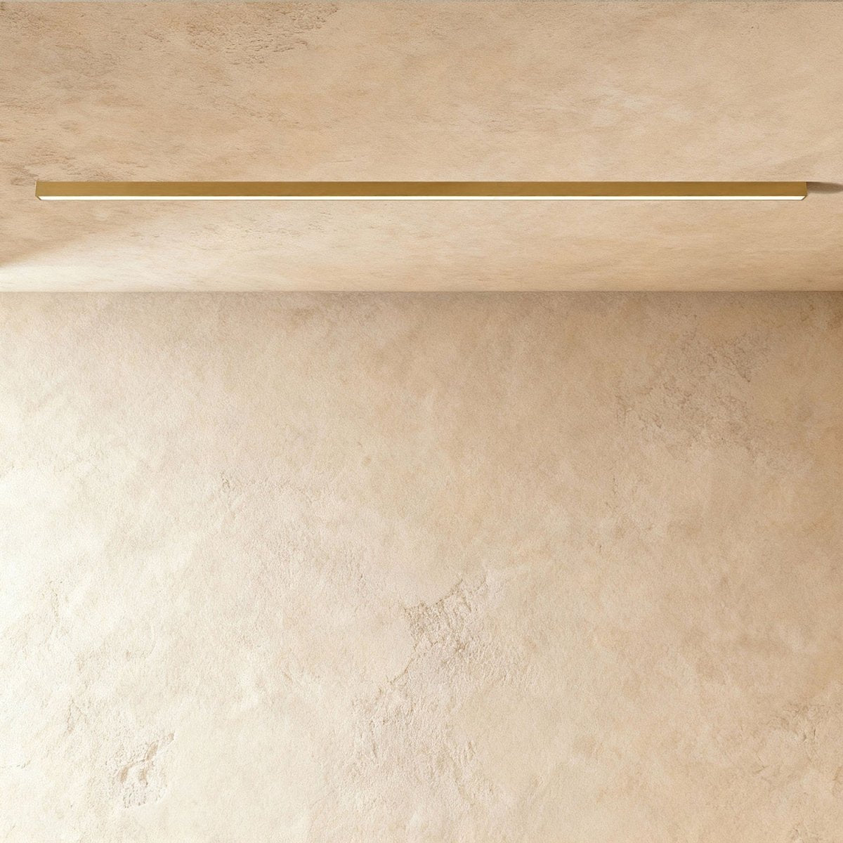 A brushed gold linear LED ceiling bar sits lit flush in a ceiling corner, this Clérachic fixture shown against a softly textured wall.