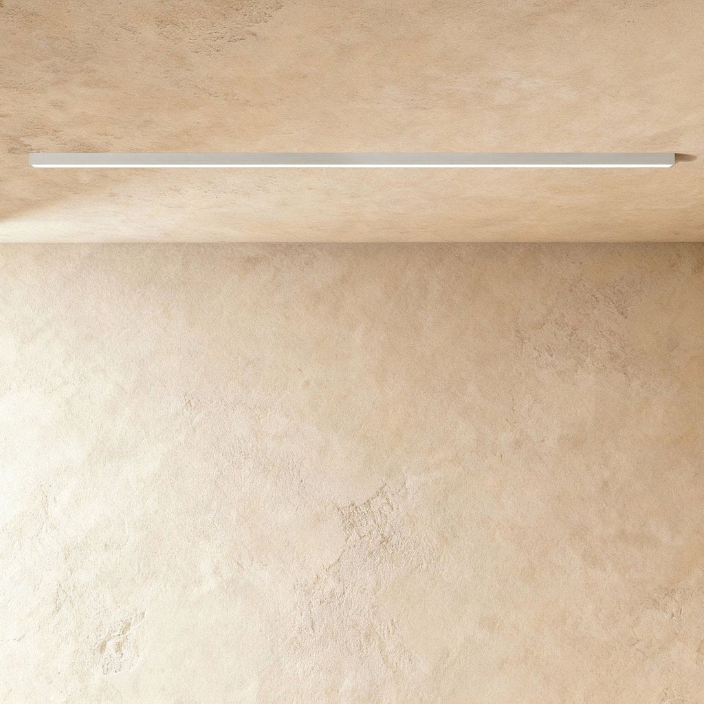 A soft white linear LED ceiling bar sits lit flush in a ceiling corner, this Clérachic fixture shown against a softly textured wall.