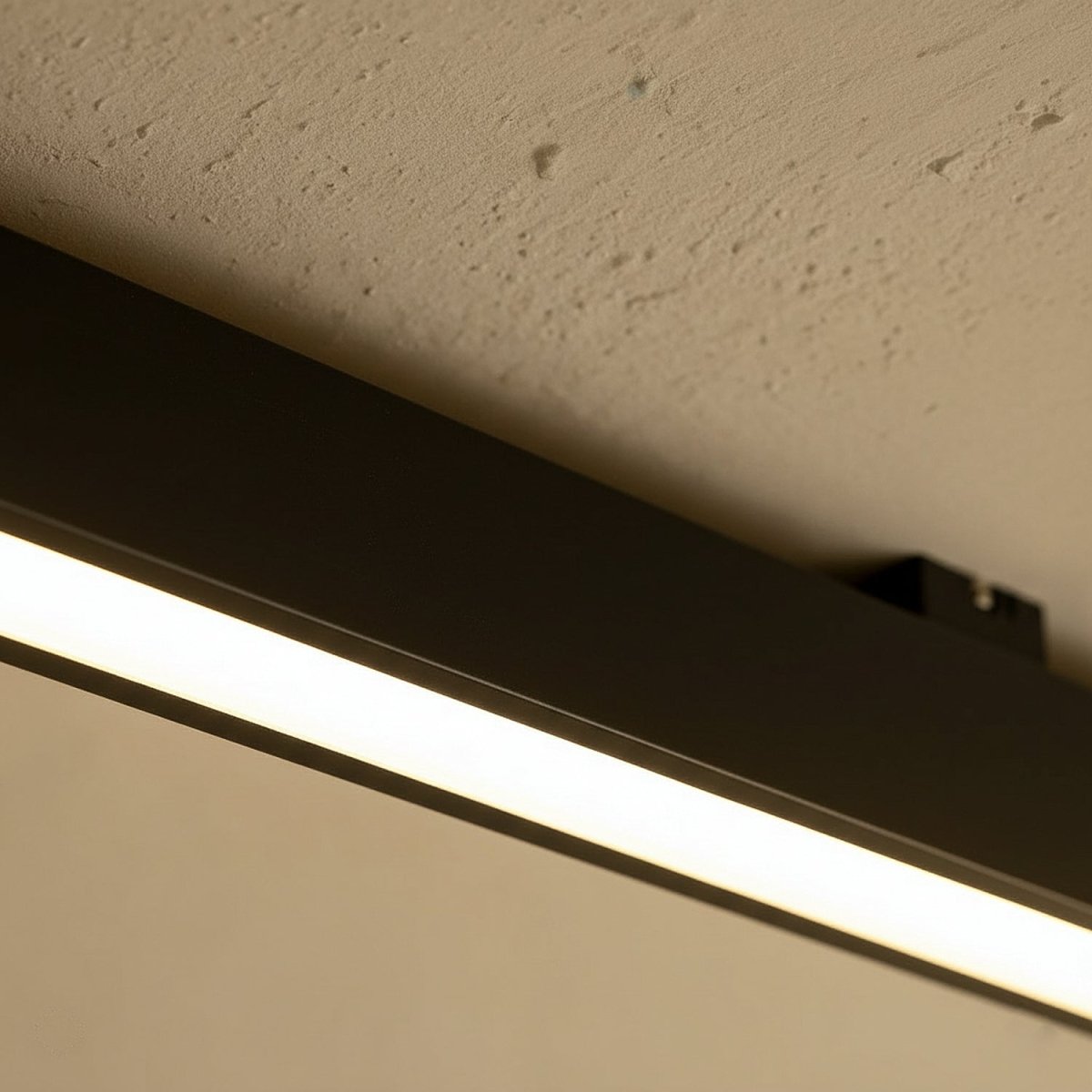 A close view of a black linear LED bar's mounting bracket against a textured stucco ceiling, this Clérachic fixture lit along its edge.