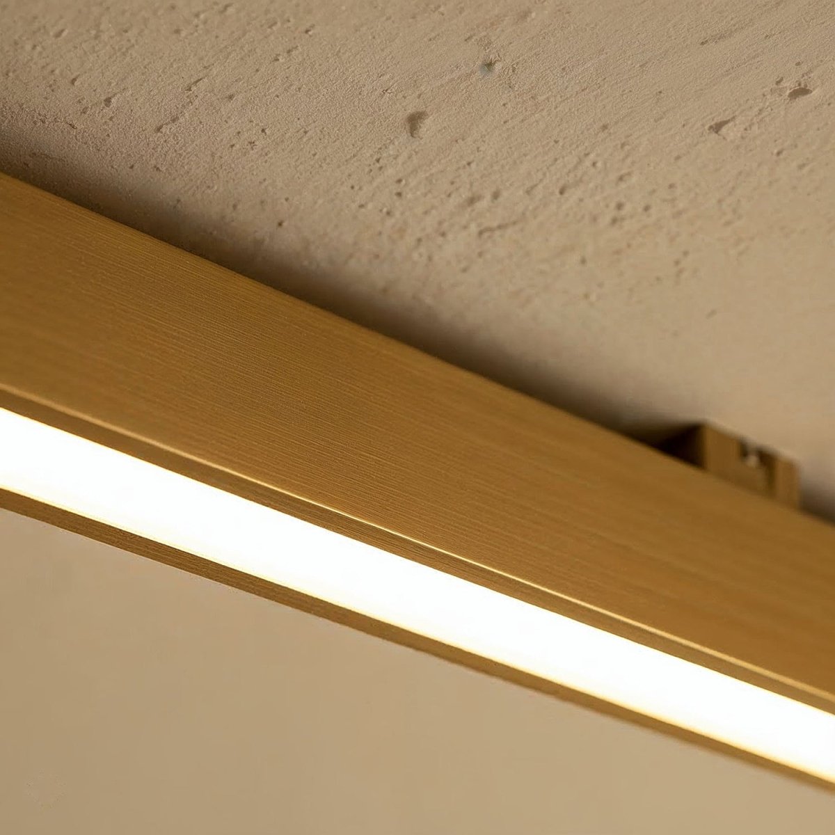 A close view of a brushed gold linear LED bar's mounting clip against a textured stucco ceiling, this Clérachic fixture lit along its edge.