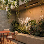 A black linear LED bar with floodlight lenses lights a shaded concrete patio hung with trailing plants, this Clérachic fixture wide view.