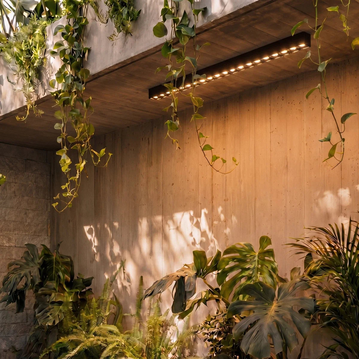 A closer view of a black linear LED bar's floodlight lenses lighting a concrete patio wall through hanging vines, a Clérachic fixture.