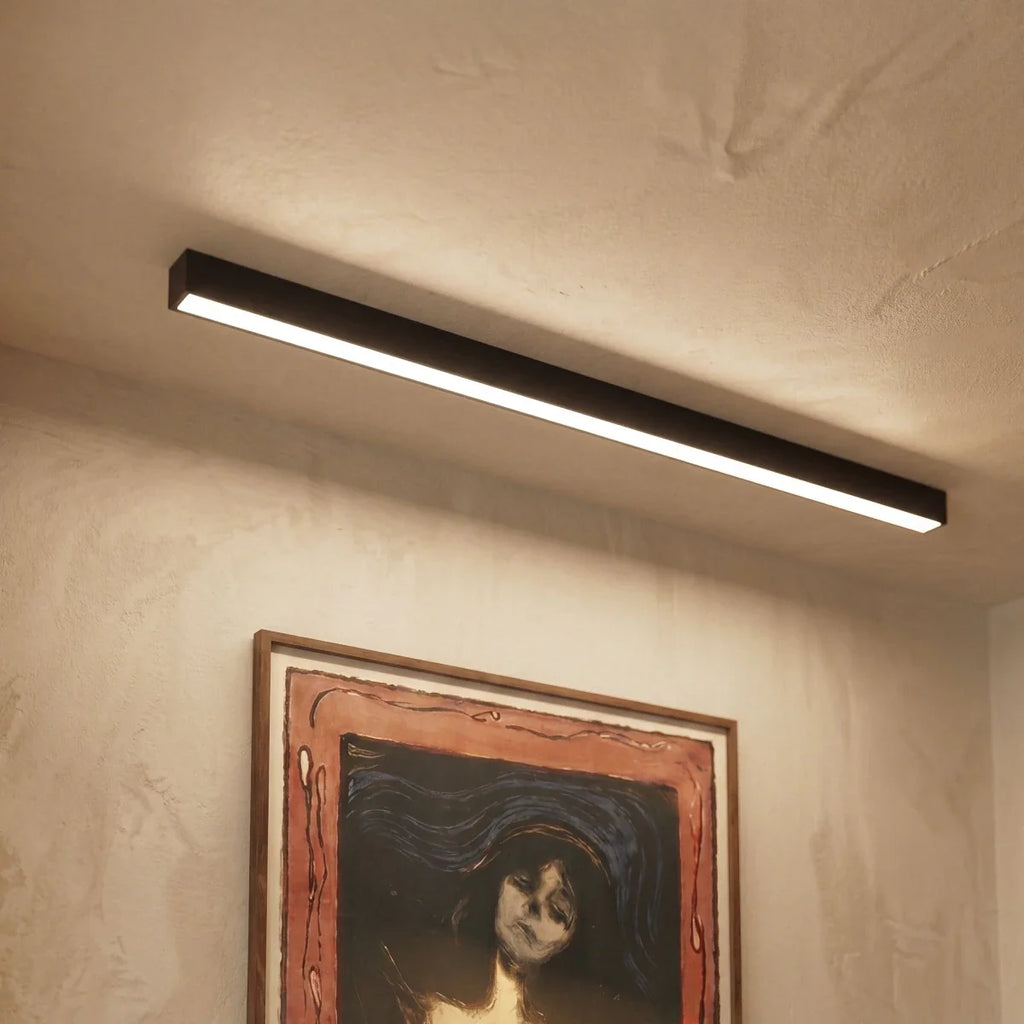 A black linear LED bar glows above a textured wall hung with a framed print, this Clérachic fixture seen close in an entryway setting.