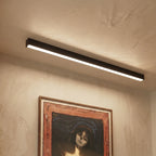 A black linear LED bar glows above a textured wall hung with a framed print, this Clérachic fixture seen close in an entryway setting.