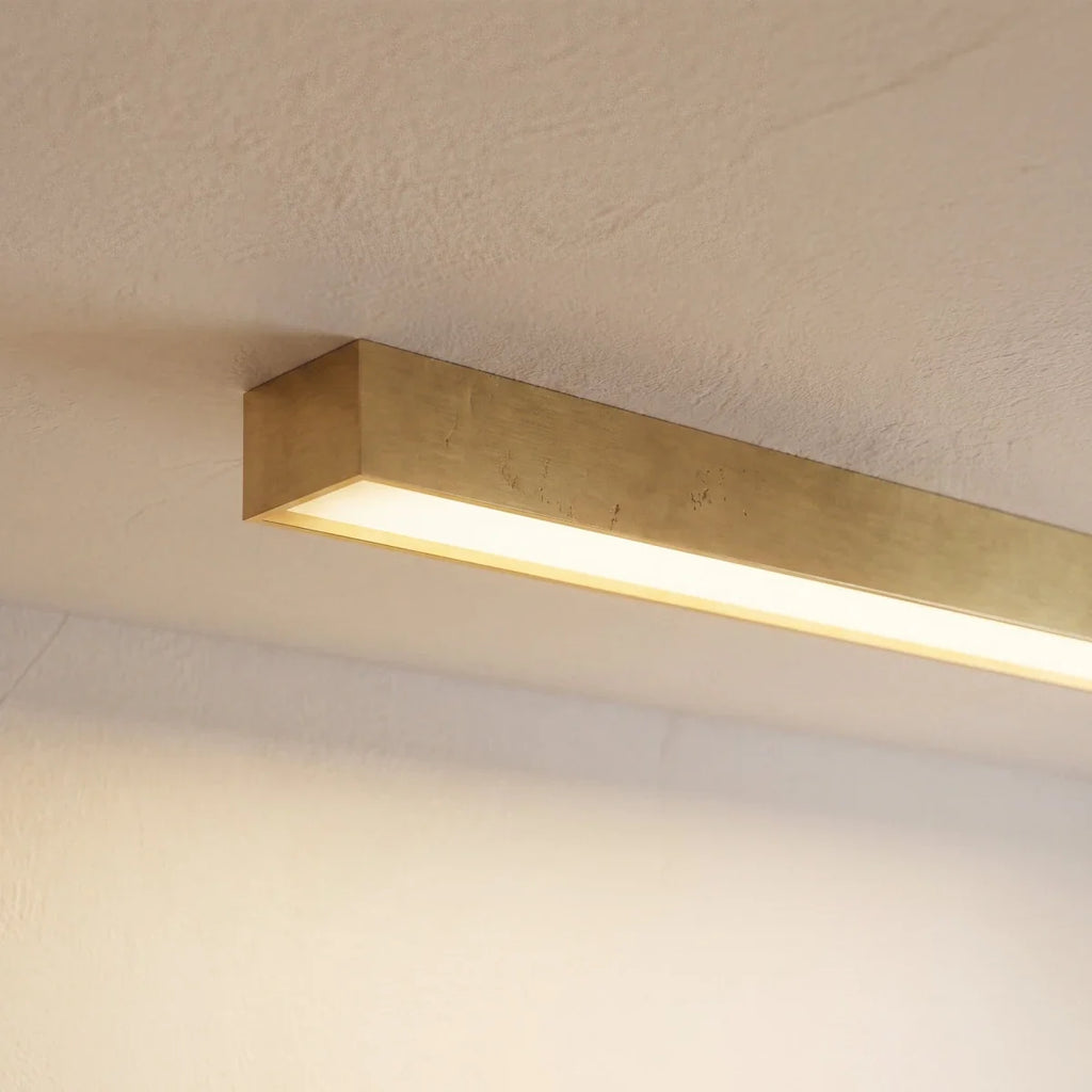 A close angled view of a brushed gold linear LED bar's lit end against a plain wall, this Clérachic fixture showing its metal finish.