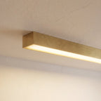 A close angled view of a brushed gold linear LED bar's lit end against a plain wall, this Clérachic fixture showing its metal finish.