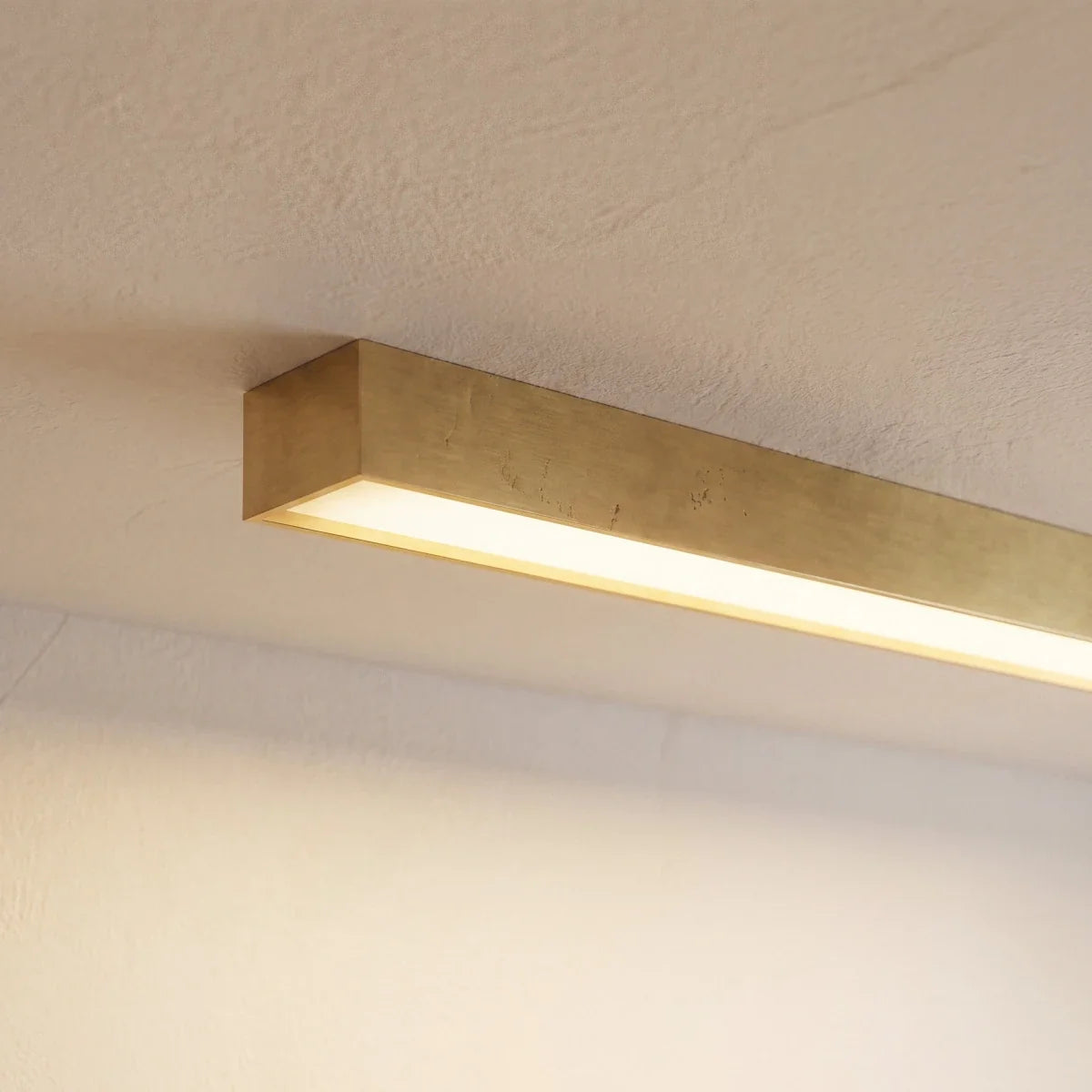 A close angled view of a brushed gold linear LED bar's lit end against a plain wall, this Clérachic fixture showing its metal finish.
