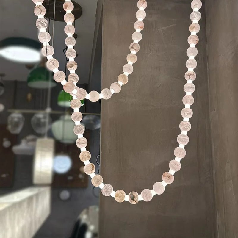 The Clérachic alabaster bead chandelier hangs in a showroom setting, reflected beside pendant lamps in the background.