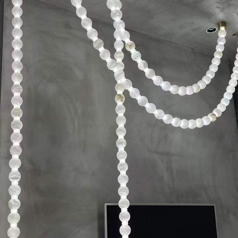 Multiple lit strands of the Clérachic alabaster bead chandelier hang against a concrete wall near a wall-mounted screen.