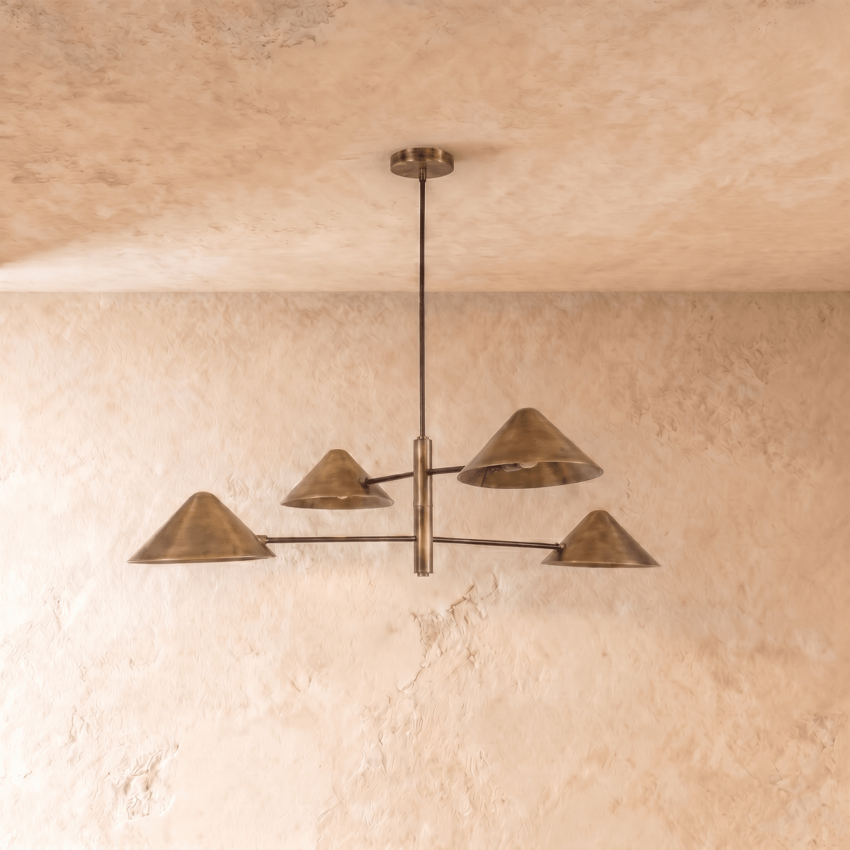 Clérachic four-arm linear chandelier in vintage brass, cone shades angled outward under a softly textured ceiling.