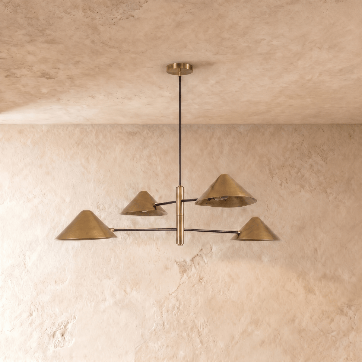 Clérachic four-light linear chandelier in satin brass, cone shades glowing warm against a plaster ceiling.