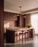 Clérachic linear chandelier in brass hanging over a stone kitchen island with walnut cabinetry and bar stools.