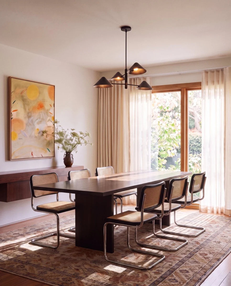 Clérachic linear chandelier in matte black hanging over a dark wood dining table with sheer curtains behind it.