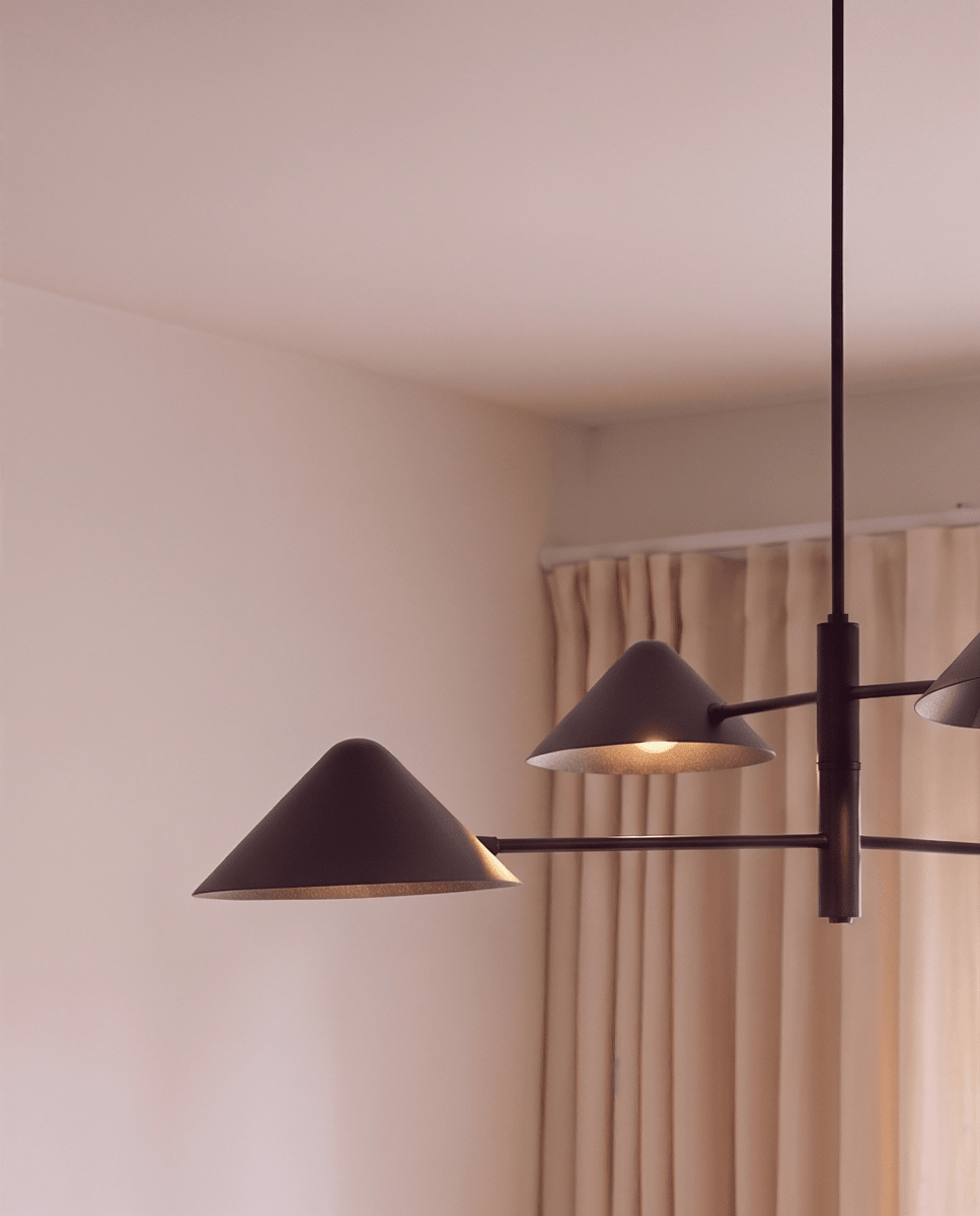 Clérachic linear chandelier detail in matte black, cone shades and crossed arms shown against a curtained window.