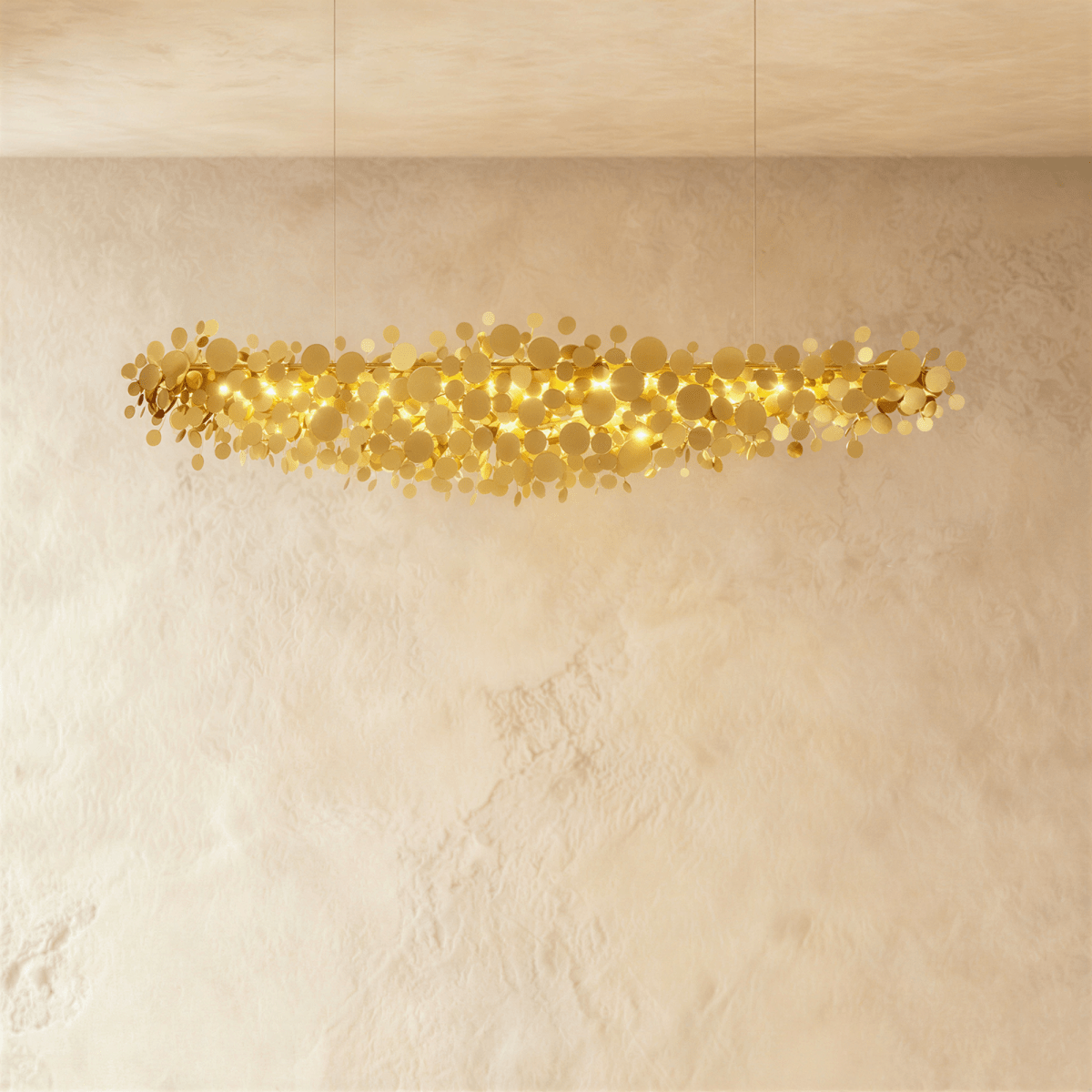 Clérachic linear chandelier of gold-finish metal discs hung from two fine cables, glowing warmly against a limewash wall.