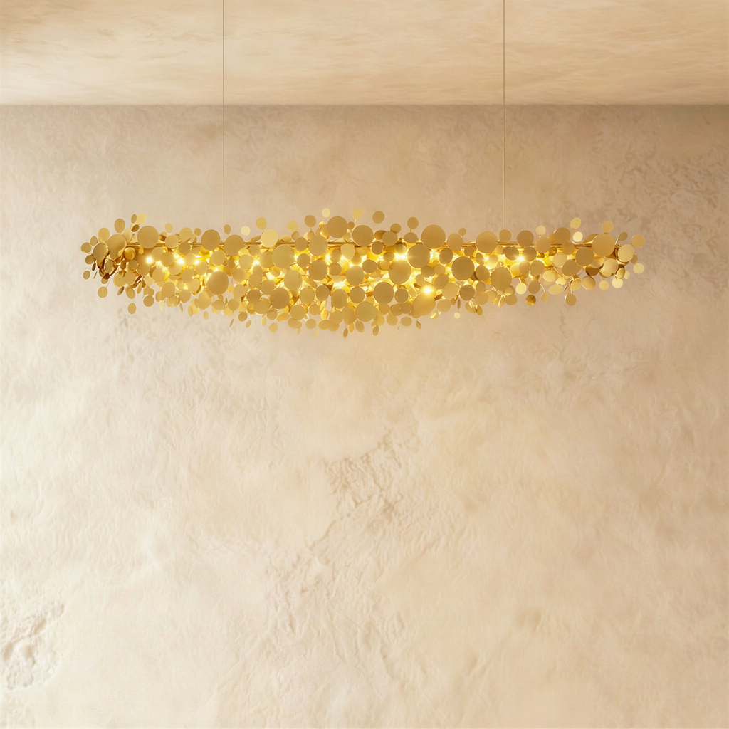 Clérachic long gold disc chandelier lit warmly, hanging from two cables in front of a softly textured plaster wall.