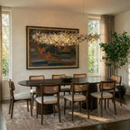 Clérachic silver disc chandelier above an oval dining table with cane-back chairs, in front of a large framed painting.
