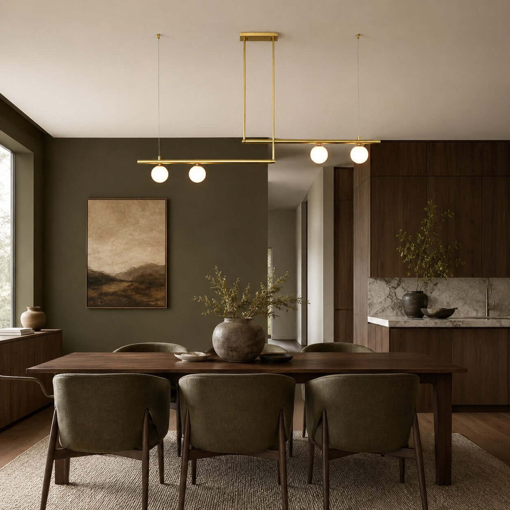 Four lit globes on two offset brass bars, from Clérachic, hang above a walnut dining table and olive-green chairs, against a dark green wall.