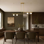 Four lit globes on two offset brass bars, from Clérachic, hang above a walnut dining table and olive-green chairs, against a dark green wall.