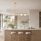 This Clérachic chandelier has one brass bar and two lit frosted globes, above a marble island with brass-legged stools and a bright garden view.