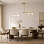 Clérachic chandelier with two brass bars at different heights, four globes glowing, above a stone dining table and cream chairs.