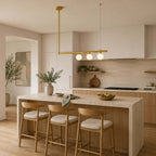 Clérachic brass chandelier with three globes lit, hung close to a hood in a kitchen with a travertine island and wooden stools.