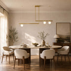 A two-bar Clérachic chandelier, its globes lit, hangs above an oval dining table with curved cream chairs and a sunlit curtain.