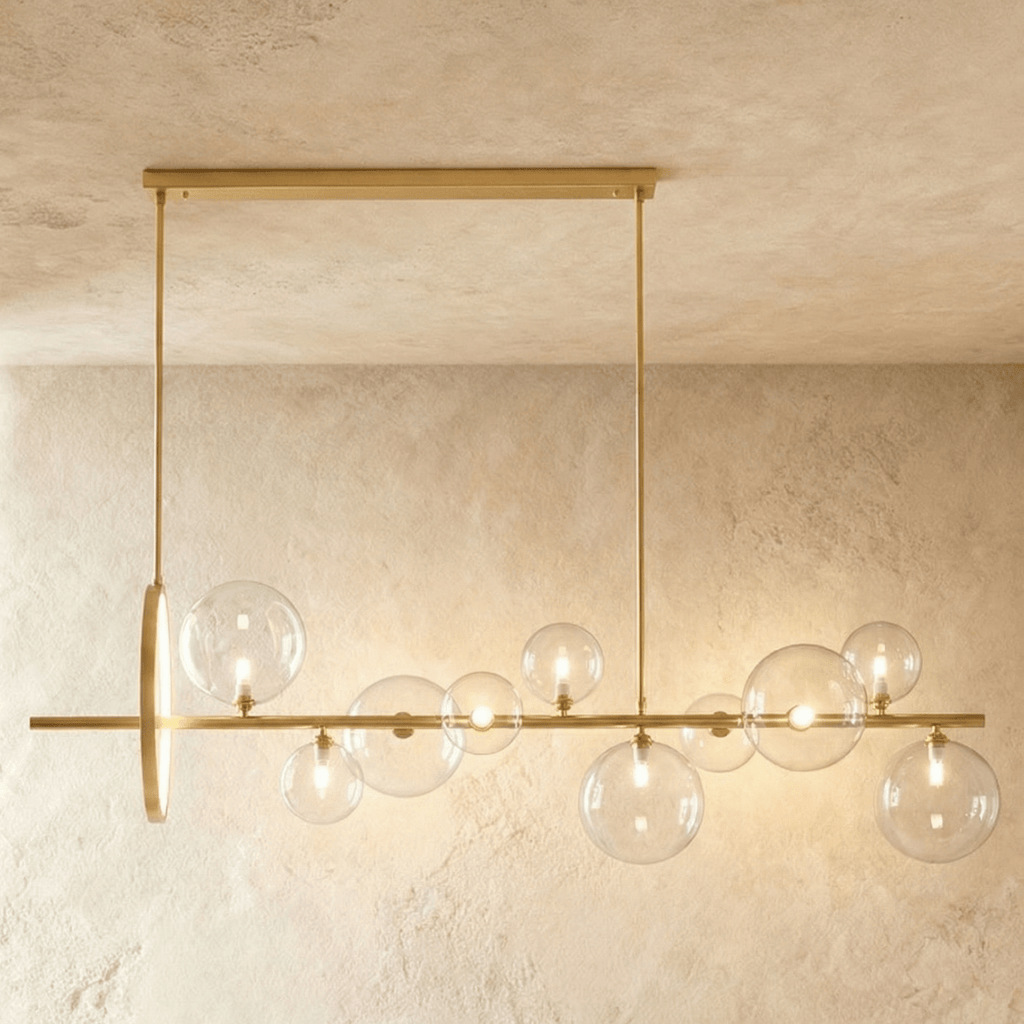 Clérachic long gold bar chandelier with ten clear glass globes of mixed sizes and a slim gold ring, hung over a plaster wall.