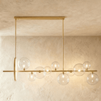 Clérachic long gold bar chandelier with ten clear glass globes of mixed sizes and a slim gold ring, hung over a plaster wall.