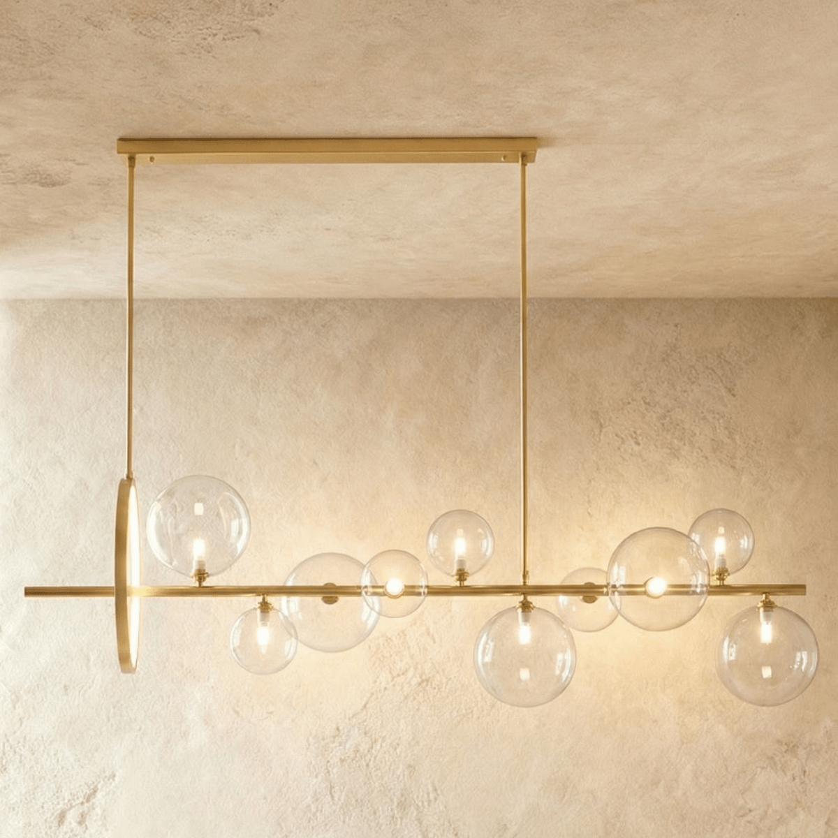 Clérachic long gold bar chandelier with ten clear glass globes of mixed sizes and a slim gold ring, hung over a plaster wall.
