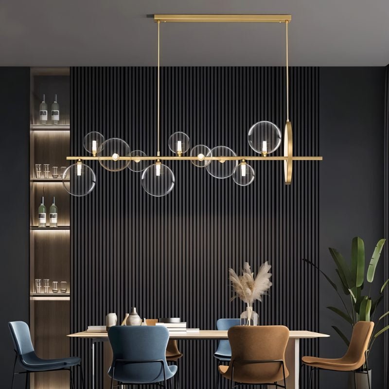 Clérachic gold bar chandelier with clear glass bubble globes above a dining table with blue and tan chairs, dark slatted wall behind.