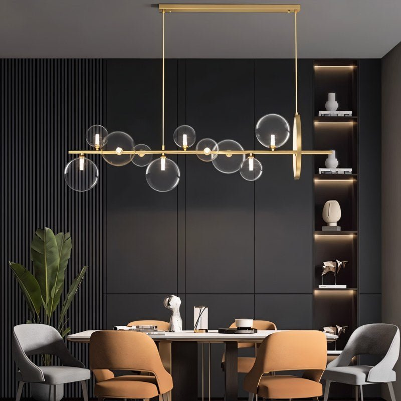 Clérachic gold bar chandelier with clear glass globes over a dining table with tan and grey chairs, dark panelled wall and shelves.