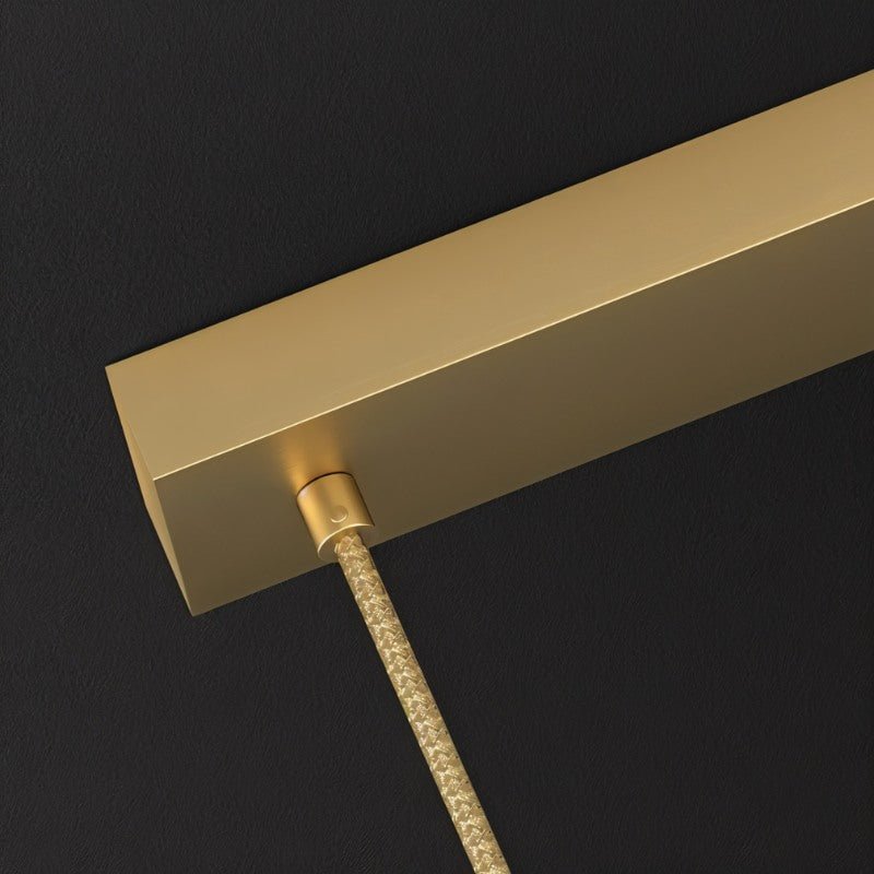 Clérachic close up of the rectangular brushed gold ceiling bar with a braided gold cord leaving it, on a dark background.