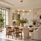 Gold vine chandelier from Clérachic lit with ceramic magnolia blossoms above a dining table with tan chairs, in a bright arched room with a large abstract painting.