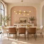 A Clérachic linear chandelier of ceramic magnolia blooms over a dining table with tan chairs, in front of a blush-pink arched alcove.