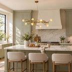 A Clérachic branch chandelier with glowing ceramic blooms above a marble kitchen island with cream stools and sage-green cabinets.