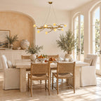 Slim Clérachic blossom chandelier above a stone dining table with woven chairs and slipcovered seats, near an arched blush alcove.