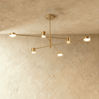 Clérachic six-head gold chandelier with slender arms and cylindrical lights, hung from a round canopy against a pale limewashed wall.