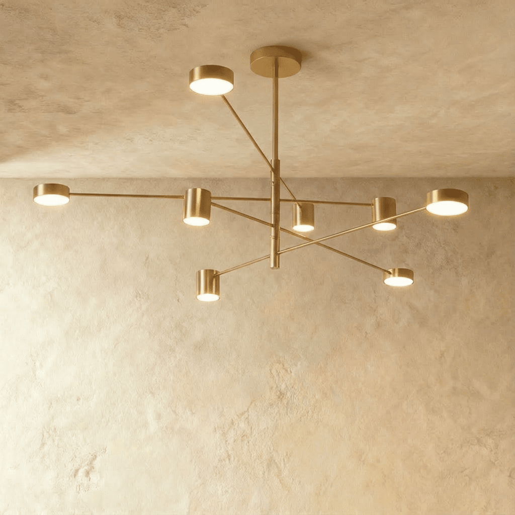 Clérachic eight-head gold chandelier with slender branching arms and cylindrical lights, seen against a pale limewashed wall and ceiling.