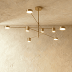 Clérachic eight-head gold chandelier with slender branching arms and cylindrical lights, seen against a pale limewashed wall and ceiling.