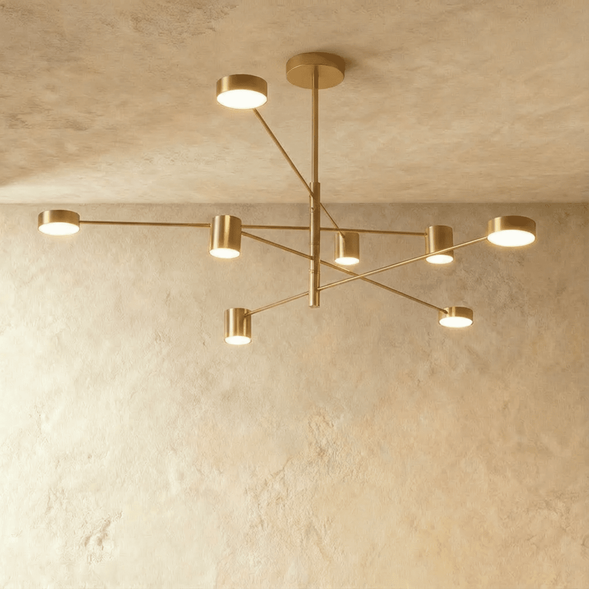 Clérachic eight-head gold chandelier with slender branching arms and cylindrical lights, seen against a pale limewashed wall and ceiling.