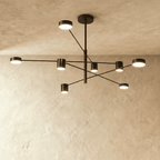 Clérachic eight-head chandelier in dark bronze-black, with slender arms and cylindrical lights, against a pale limewashed wall.