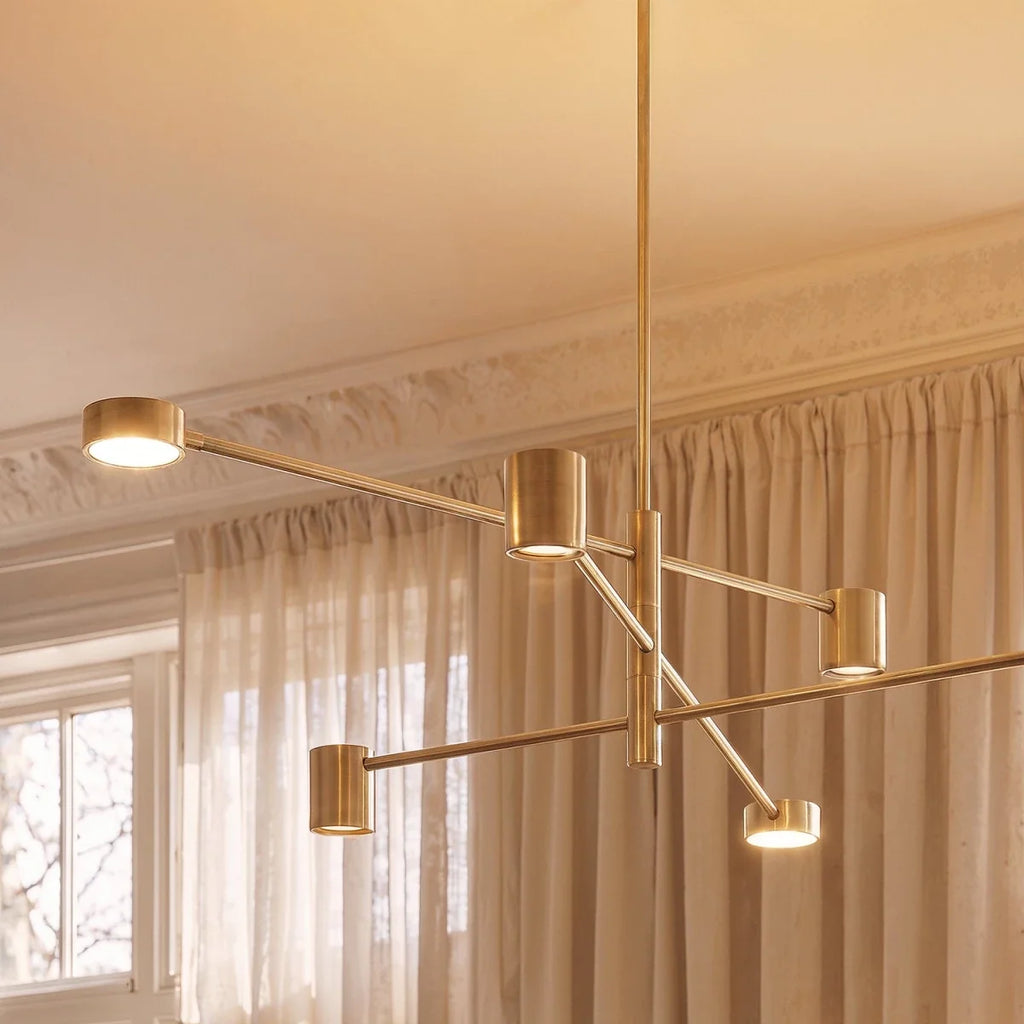 Close view of a Clérachic gold chandelier showing its slim brushed arms, cylindrical heads and warm glowing diffusers beside sheer curtains.